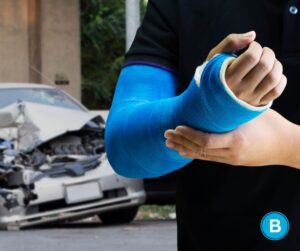 Man with injured arm in blue cast is standing in front of a wrecked car.