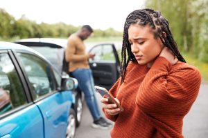 woman of color rubs back of neck while looking at phone after car accident