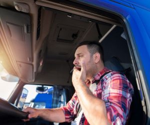 trucker yawning with hand over mouth while behind the wheel of semi truck