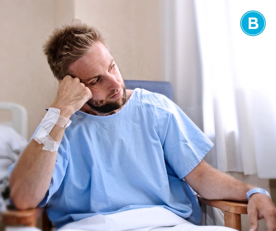 man sits in wheelchair wearing blue hospital gown and looking distraught and distant