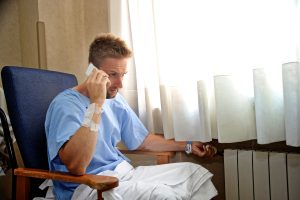man talks on phone in wheelchair sitting by hospital window. Wearing hospital gown.