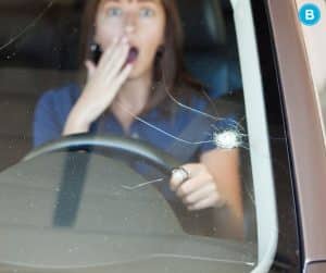 woman holds hand over mouth and looks shocked behind wheel of her car after rock hits windshield and leaves large crack in glass