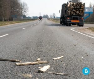 logging debris fallen off logging truck with logging truck parked on side of road in distance