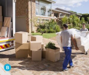 Two men loading a moving truck. They're carrying a sofa, and there are boxes stacked on the ground around them at the back of the moving truck.