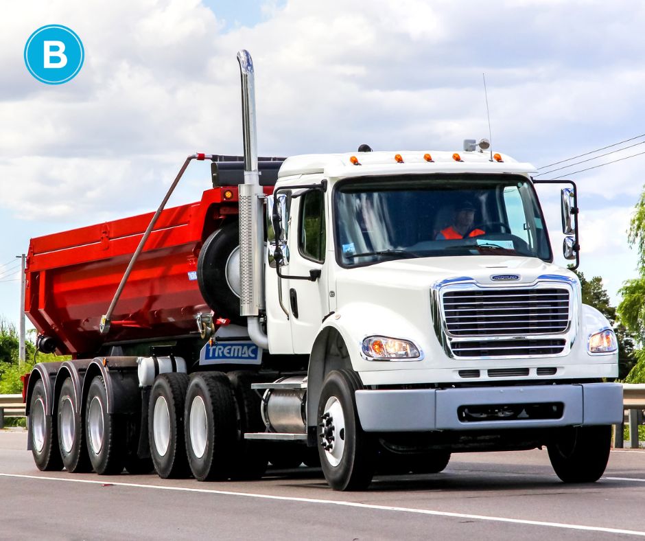 A large dump truck with a red bed and white tractor travels down a highway with blue skies.