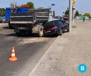 A small, dark sedan is crushed between a dump truck on the left and a light pole on the right. Traffic cones surround area.